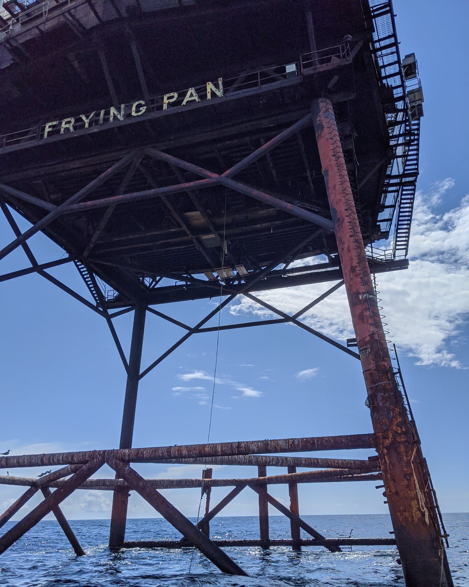 Rust-streaked steel legs and cross-bracing beneath the station's deck, seen from a boat, with the name FRYING PAN painted along the deck's edge.